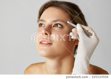 Beautiful young woman taking care after look, appearance plucking eyebrows with tweezer against grey studio background Beautiful young woman taking care after look, appearance plucking eyebrows with tweezer against grey studio background 105534504