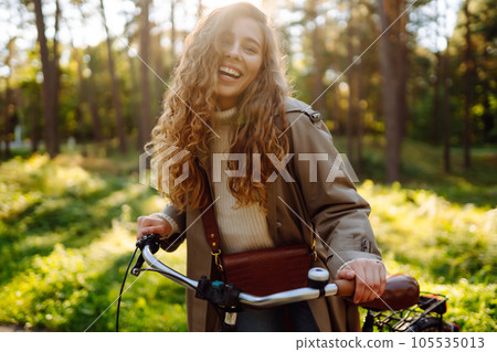 Smiling woman with curly hair in coat rides bicycle in sunny park. Lifestyle. Relax, nature concept. 105535013