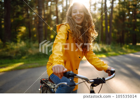 Young happy woman in yellow coat rides bicycle in sunny park. Relax, nature concept. Lifestyle. 105535014