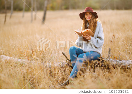 Fall picnic in nature. Young woman sitting on the blanket reading a book and smiling outdoors. Fall picnic in nature. Young woman sitting on the blanket reading a book and smiling outdoors. 105535981