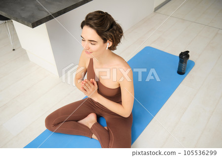 Young attractive smiling woman practicing yoga, sitting in Half Lotus exercise, Ardha Padmasana pose, working out, wearing sportswear, meditation session, indoor full length, home interior Young attractive smiling woman practicing yoga, sitting in Half Lotus exercise, Ardha Padmasana pose, working out, wearing sportswear, meditation session, indoor full length, home interior 105536299