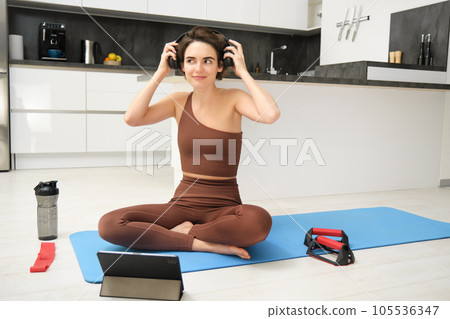 Gym at home. Young smiling woman puts on headphones in her kitchen, wearing sportswear, ready for workout, follows fitness instructor app online video on tablet, sits with exercise equipment 105536347
