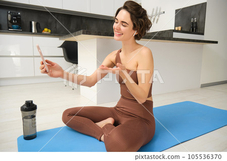 Portrait of happy smiling, relaxed fitness instructor, girl video chats, looks at smartphone, records workout blog, yoga online, sits on sports mat in kitchen, does pilates from home 105536370