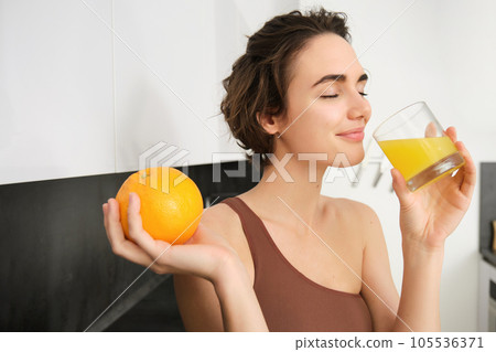 Healthy lifestyle and sport. Beautiful smiling woman, drinking fresh orange juice and holding fruit in her hand, enjoying her vitamin drink after workout at home, standing in kitchen 105536371