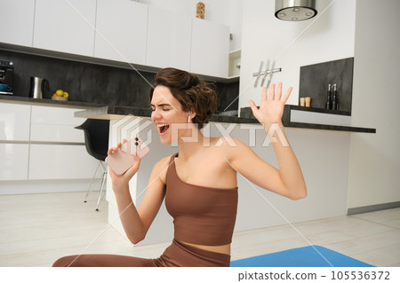 Portrait of brunette girl, does workout at home, sings in smartphone, listens music in wireless headphones while doing exercises, fitness training on kitchen floor, sport and healthy lifestyle 105536372