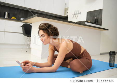 Young fitness girl sitting in her kitchen on yoga mat and scrolling exercise videos on mobile phone, does stretching workout at home, wears sportswear. Online gym application 105536380