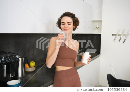 Portrait of fitness girl, healthy woman standing in kitchen with pills, takes vitamins with glass of water for healthy wellbeing. Sportswoman holds food supplements 105536384