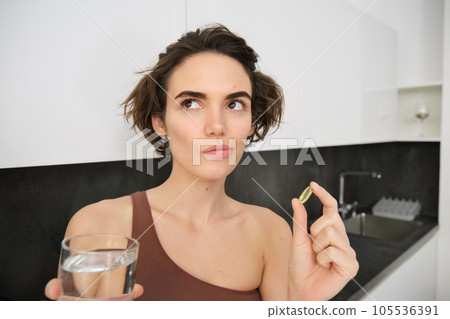 Close up portrait of sportswoman in activewear, holding omega-3, vitamin c, d and b in her hand, taking dietary supplement with drinking glass of water. Healthy lifestyle and sport concept 105536391