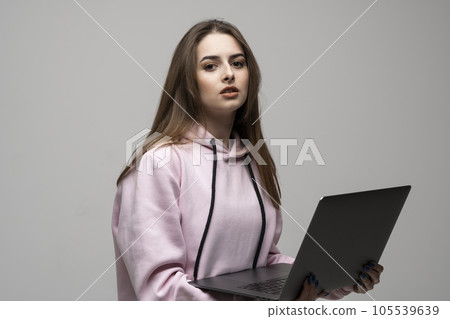 Attractive brunette woman 20s wearing pink hoodie standing working on laptop pc computer isolated on grey wall background studio portrait. Attractive brunette woman 20s wearing pink hoodie standing working on laptop pc computer isolated on grey wall background studio portrait. 105539639