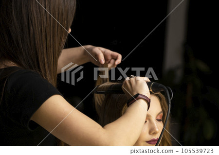 The work of the hair stylist, hairdresser with hairstyles for a beautiful young woman client. Stylist curls hair with clipless curling iron. 105539723