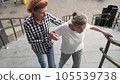Senior woman and her middle-aged daughter walk in courtyard of high-rise building. Nurse helps an old pensioner climb on stairs outside. Caregiver helping, assistance and support the elderly people 105539738