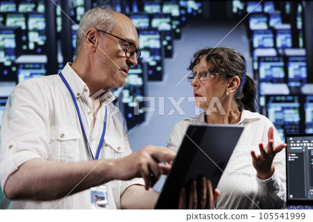 Elderly servicemen running analysis on supercomputers helping businesses manage databases, host websites and store files. Meticulous repairmen auditing high tech server farm electronics 105541999