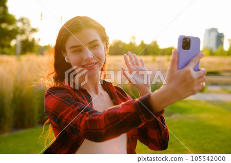 A portrait of a happy red-haired young blogger in a plaid shirt warmly greets her followers, recording a captivating story using her mobile phone at sunset A portrait of a happy red-haired young blogger in a plaid shirt warmly greets her followers, recording a captivating story using her mobile phone at sunset 105542000
