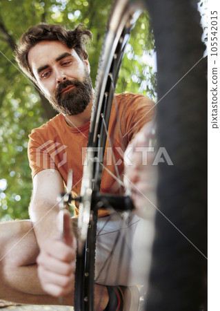 Committed healthy and active man outdoor showing dedication in adjusting and securing bicycle parts for recreational summer cycling. Image showing young man fixing bike wheel, summer activity. Committed healthy and active man outdoor showing dedication in adjusting and securing bicycle parts for recreational summer cycling. Image showing young man fixing bike wheel, summer activity. 105542015