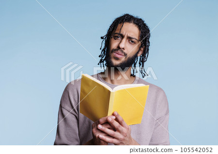 Confused arab man reading paperback book and looking at camera with puzzled facial expression. Young questioned person holding open softcover textbook and learning portrait Confused arab man reading paperback book and looking at camera with puzzled facial expression. Young questioned person holding open softcover textbook and learning portrait 105542052