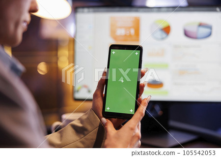 Entrepreneur holding mobile phone with green screen chroma key mock up display, working late at night in startup office. Executive manager analyzing marketing graphs report. Business concept 105542053