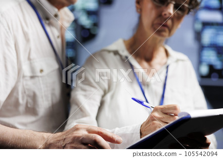 Aged colleagues monitoring server rows, optimizing them for data processing requirements of AI applications. Licensed experts working in high tech facility used for machine learning 105542094