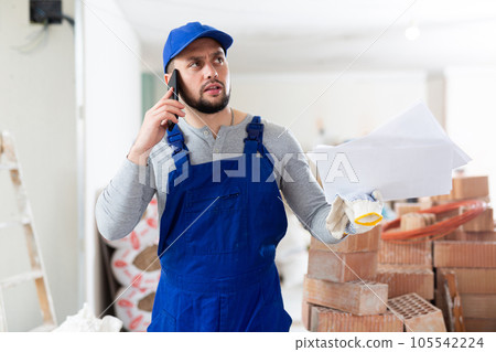 Master in blue overalls talking on mobile phone, discussing documents in renovated building Master in blue overalls talking on mobile phone, discussing documents in renovated building 105542224