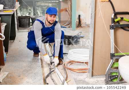 Qualified builder in blue workwear plans to make warm floor, prepares plastic pipes 105542249