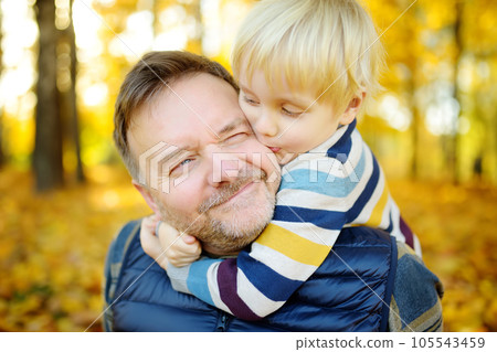 Little boy embraces his mature father and kissing him during stroll at sunny autumn park Little boy embraces his mature father and kissing him during stroll at sunny autumn park 105543459