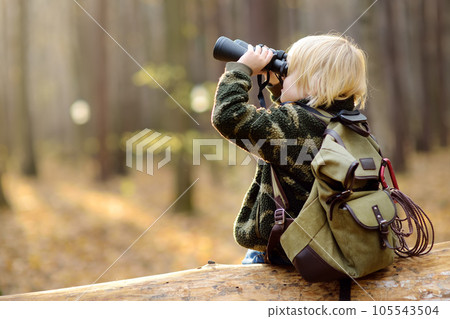 Little boy scout with binoculars during hiking in autumn forest. Child is sitting on large fallen tree and looking through a binoculars. Little boy scout with binoculars during hiking in autumn forest. Child is sitting on large fallen tree and looking through a binoculars. 105543504