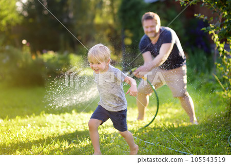 Funny little boy with his father playing with garden hose in sunny backyard. Preschooler child having fun with spray of water. Funny little boy with his father playing with garden hose in sunny backyard. Preschooler child having fun with spray of water. 105543519
