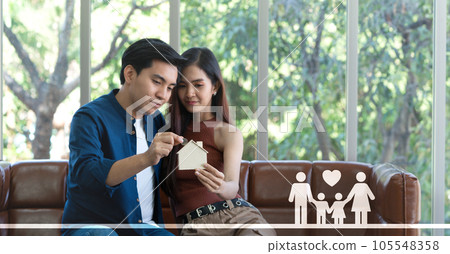 Young asian couple sits on a cozy couch, putting silver coin into a wooden house-shaped storing coin box , hinting at dreams of an idyllic future. 105548358
