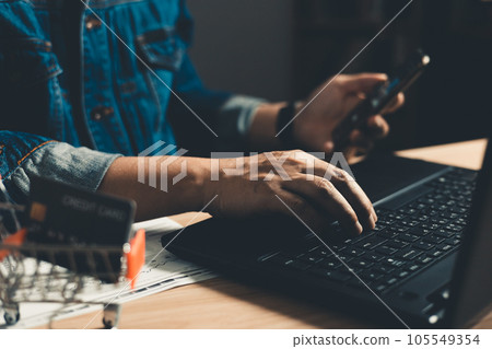 Man using smartphone and computer laptop. which is payment online, sits on the chair in the office at home on a desktop. The concept of finance and online shopping. Man using smartphone and computer laptop. which is payment online, sits on the chair in the office at home on a desktop. The concept of finance and online shopping. 105549354