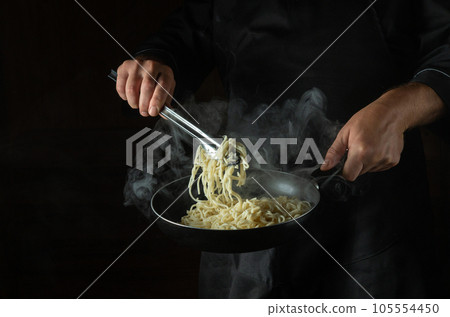 The chef fries spaghetti in a hot pan with steam. Space for advertising on a black background. Italian national dish 105554450