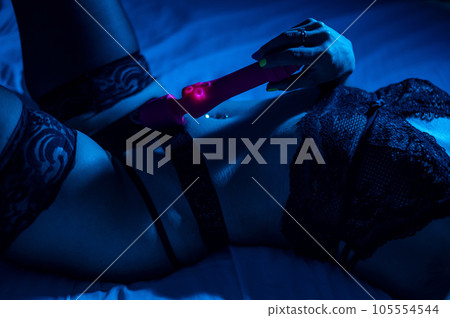 A woman in black lace lingerie lies on a bed in the dark and holds a vibrator. A woman in black lace lingerie lies on a bed in the dark and holds a vibrator. 105554544