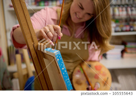 Focus on hand of female painter holding paint brush, drawing picture on canvas, standing at wooden easel in her workshop 105555643