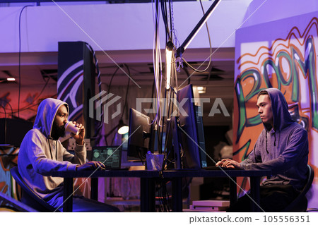 Hackers breaking law and doing illegal activity on computer late at night. Two young diverse criminals drinking coffee while programming malicious software in dark hideout place Hackers breaking law and doing illegal activity on computer late at night. Two young diverse criminals drinking coffee while programming malicious software in dark hideout place 105556351