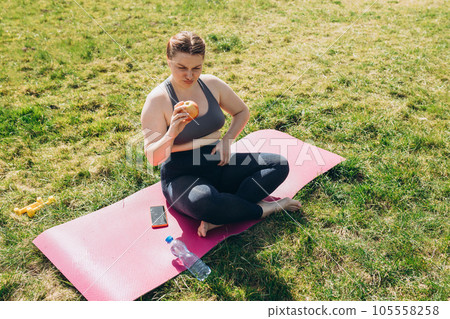 Young angry woman wearing sportswear eating apple outdoors. Fat woman dieting, fitness and healthy food on nature backgrond 105558258