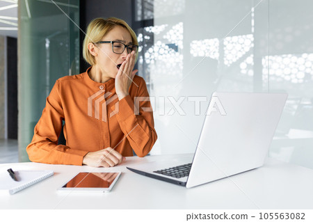 Boring and boring routine work in office, blonde business woman yawning sitting at desk, female worker tired using laptop at workplace. 105563082