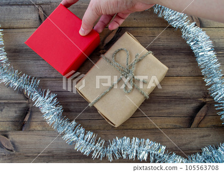 New Year gifts. A gift in craft paper, a hand holds a gift in a red box, tinsel on a wooden background. New Year gifts. A gift in craft paper, a hand holds a gift in a red box, tinsel on a wooden background. 105563708