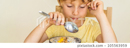 Happy boy sitting and eating a fresh smoothie bowl with mango, dragon fruit, granola and chia seeds BANNER, LONG FORMAT 105563959