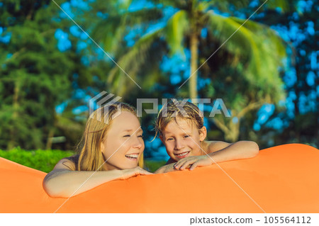 Summer lifestyle portrait of mother and son sitting on the orange inflatable sofa on the beach of tropical island. Relaxing and enjoying life on air bed Summer lifestyle portrait of mother and son sitting on the orange inflatable sofa on the beach of tropical island. Relaxing and enjoying life on air bed 105564112
