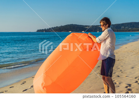 Summer lifestyle portrait of man inflates an inflatable orange sofa on the beach of tropical island. Relaxing and enjoying life on air bed Summer lifestyle portrait of man inflates an inflatable orange sofa on the beach of tropical island. Relaxing and enjoying life on air bed 105564160