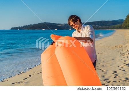 Summer lifestyle portrait of man inflates an inflatable orange sofa on the beach of tropical island. Relaxing and enjoying life on air bed 105564162