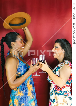 Two female multiracial friends smiling, dancing and drinking wine 105565640