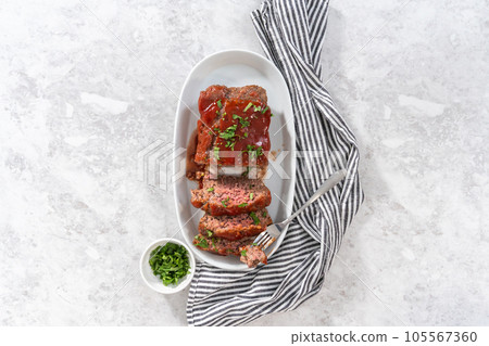 Flat lay. Slicing classic meatloaf with a sweet glaze on a white serving plate 105567360