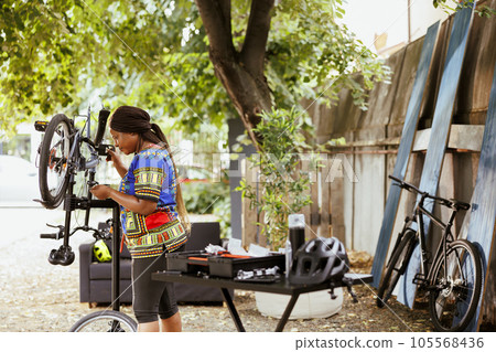 Active african american female cyclist surrounded by professional equipment to maintain bike gear and ensuring peak performance for outdoor cycling. Summer activity of servicing bicycle. Active african american female cyclist surrounded by professional equipment to maintain bike gear and ensuring peak performance for outdoor cycling. Summer activity of servicing bicycle. 105568436