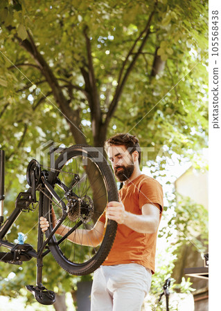 Image showing enthusiastic sporty caucasian man gripping and examining damaged dismantled bicycle wheel for repair. Healthy young active male cyclist reattaching bike tire rubber. Image showing enthusiastic sporty caucasian man gripping and examining damaged dismantled bicycle wheel for repair. Healthy young active male cyclist reattaching bike tire rubber. 105568438