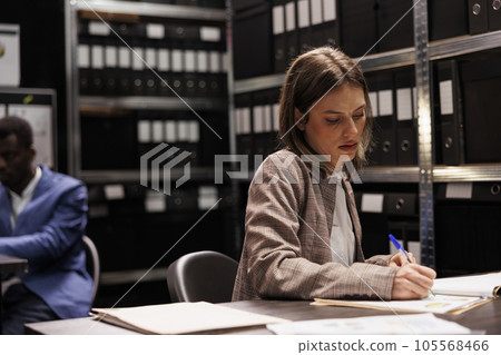 Businesswoman writing corporate report, working late at night at administrative record, analyzing bookkeeping documents in arhive room. Depository worker checking accountancy research Businesswoman writing corporate report, working late at night at administrative record, analyzing bookkeeping documents in arhive room. Depository worker checking accountancy research 105568466