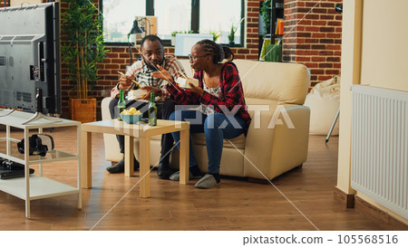 Smiling couple eating asian food from takeaway delivery, serving noodles with chopsticks and watching action film on tv. Young people in relationship enjoying movie and meal with beer. Smiling couple eating asian food from takeaway delivery, serving noodles with chopsticks and watching action film on tv. Young people in relationship enjoying movie and meal with beer. 105568516
