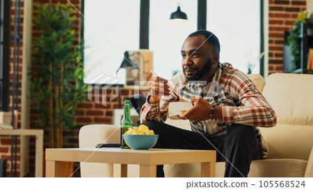 Happy guy eating noodles with chopsticks at home, enjoying asian food from delivery. Young adult relaxing with bottle of beer and takeaway meal, laughing at favorite tv show. Handheld shot. Happy guy eating noodles with chopsticks at home, enjoying asian food from delivery. Young adult relaxing with bottle of beer and takeaway meal, laughing at favorite tv show. Handheld shot. 105568524