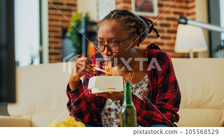 Female person using chopsticks to eat asian food, laughing at favorite film on television. Young woman eating noodles in delivery box and drinking beer, having fun at television. Handheld shot. 105568529