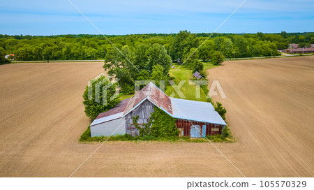 Old rusty barn in center with empty dirt farmland on three sides and a lush green forest behind aerial 105570329