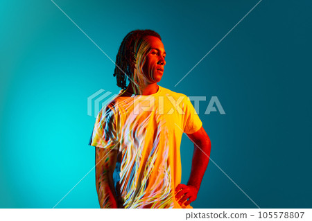 Portrait of young african man with dreads standing against blue, cyan background in neon light. Light reflection on body Portrait of young african man with dreads standing against blue, cyan background in neon light. Light reflection on body 105578807