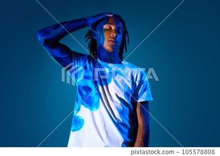 Creative portrait of african young man with dread with light reflection on body standing against blue background in neon light 105578808